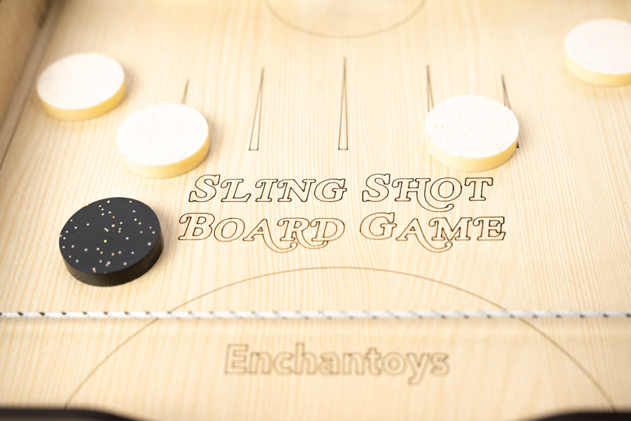 Enchantoys wooden Slingshot Board Game featuring engraved markings, black and white pucks, and tension cords on each side. A fun and competitive two-player game using flick mechanics.