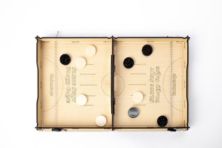 Wooden Slingshot Board Game by Enchantoys with black and white pucks placed across two playing fields. The board features a central slot and elastic bands for flick-based gameplay.